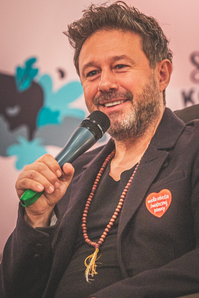 Zdjęcie. Uśmiechnięty mężczyzna w czarnym podkoszulce i marynarce mówi do mikrofonu. To Andrzej Piaseczny.