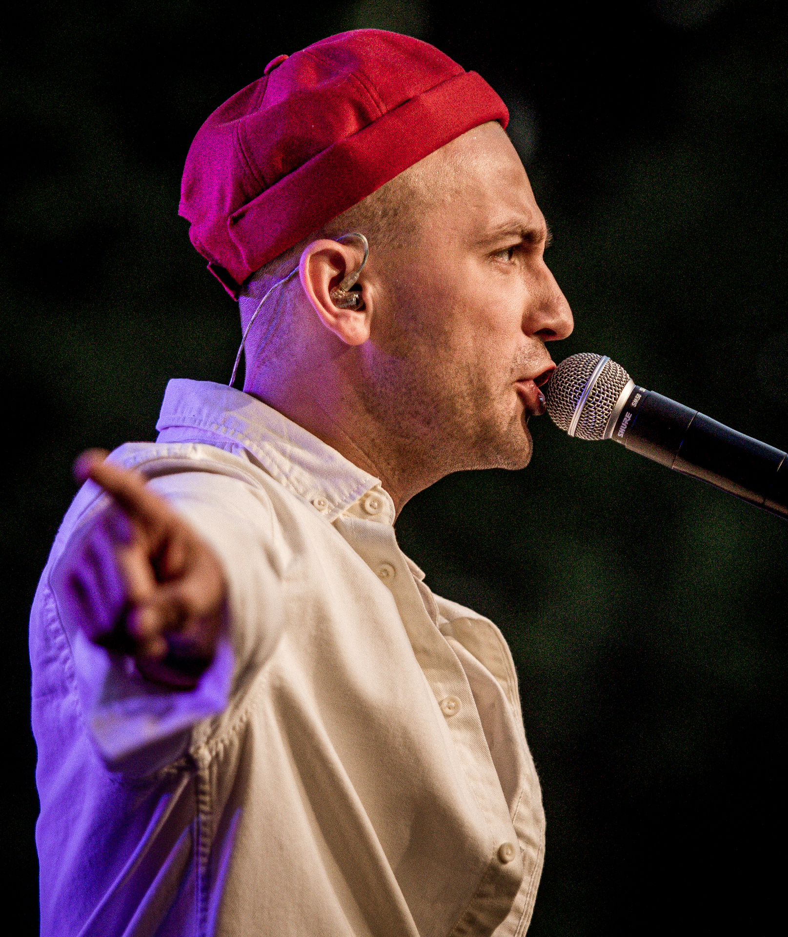 Zdjęcie. Mężczyzna w białej koszuli i czerwonej czapce na głowie śpiewa do mikrofonu. To Arek Kłusowski.