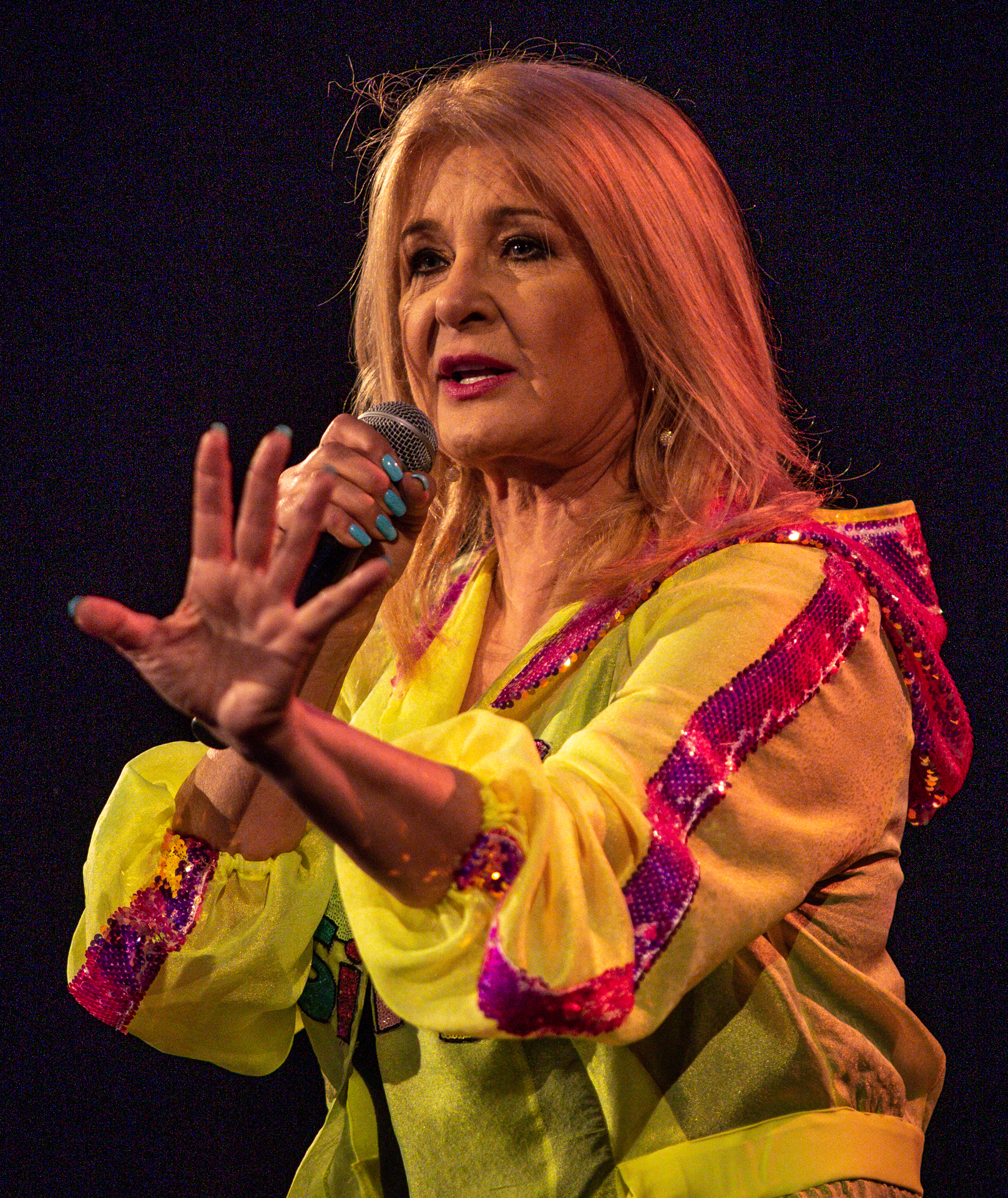 Zdjęcie. Kobieta w żółtej szyfonowej bluzie z kapturem z błyszczącymi różowymi lampasami śpiewa do mikrofonu. Ma proste blond włosy poniżej ramion. To Majka Jeżowska.