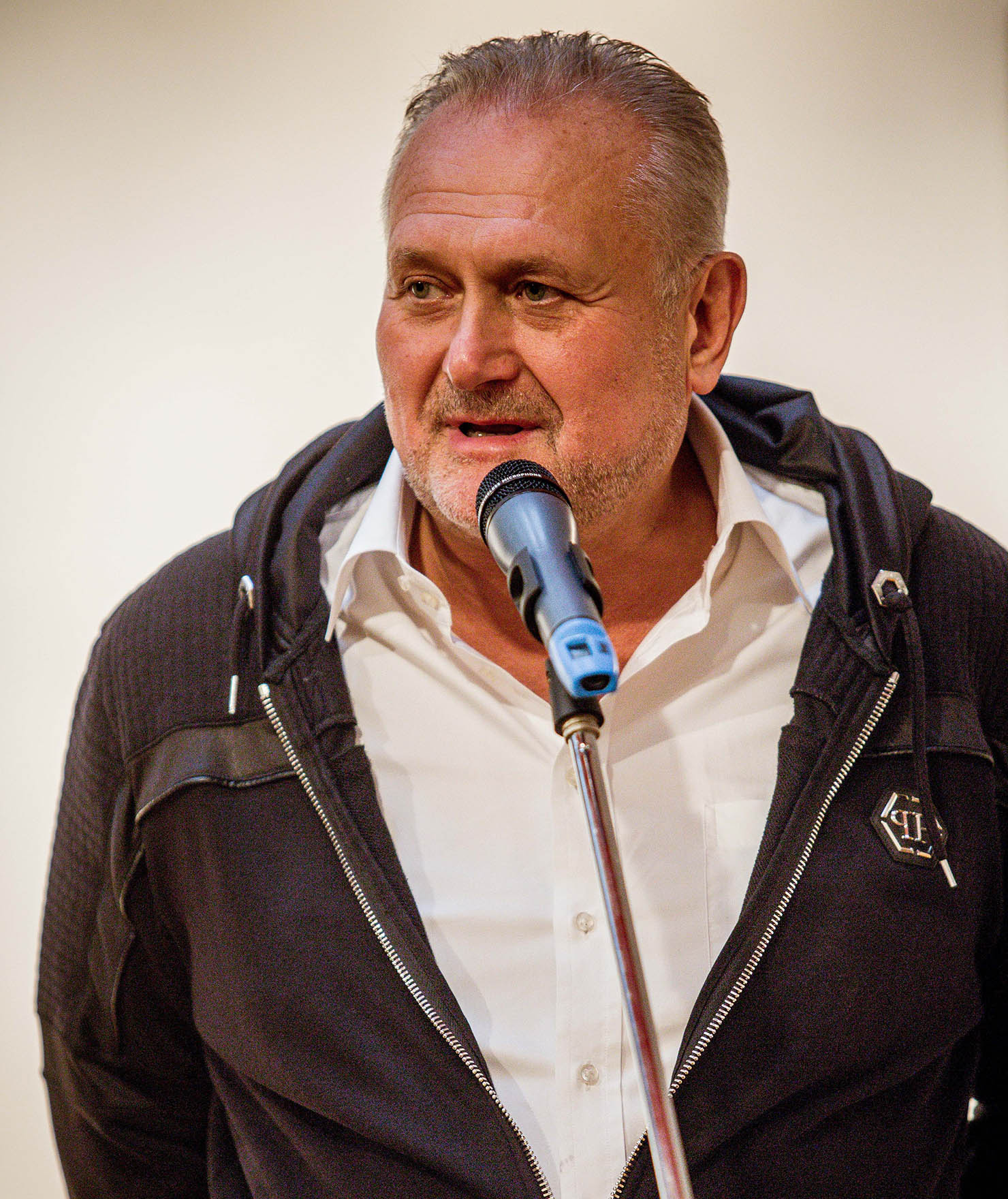 Zdjęcie. Postawny mężczyzna w białej koszuli i ciemnej bluzie z siwymi włosami i zakolami mówi do mikrofonu. To malarz Paweł Kowalewski.