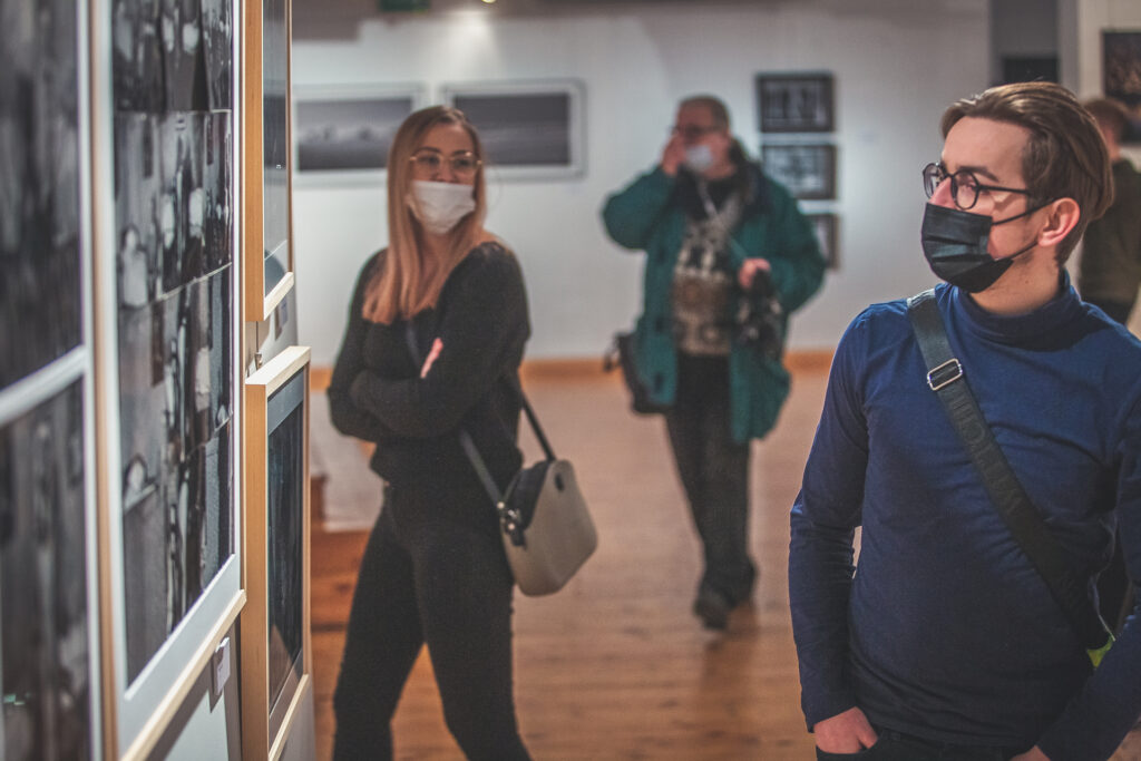 Zdjęcie. Młody mężczyzna przygląda się fotografiom wiszącym na ścianie. W tle inni goście wernisażu wystawy w Galerii Winda.