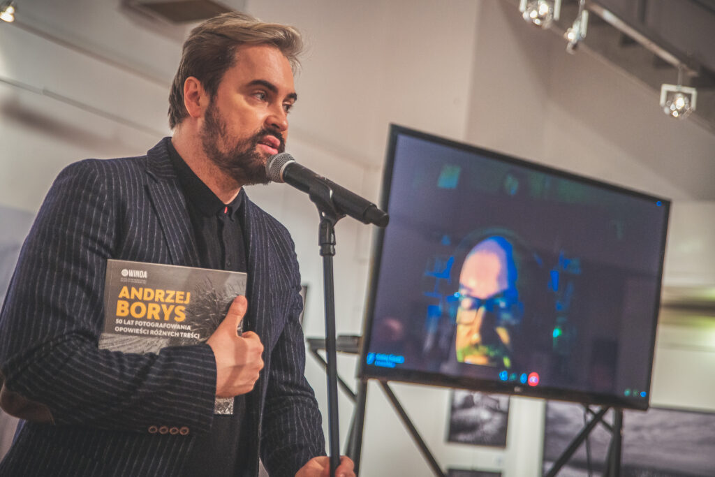 Zdjęcie. Szef Galerii Winda Artur Wijata z katalogiem wystawy fotografii Andrezja Borysa mówi do mikrofonu. W tle duży ekran, na którym widać artystę ze słuchawkami na uszach.