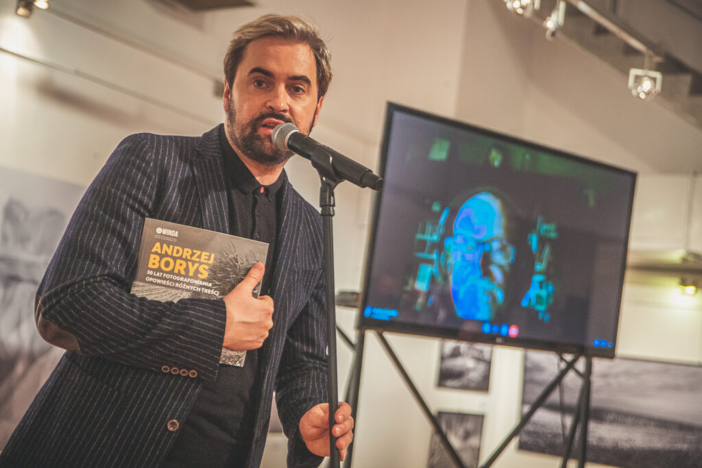 Zdjęcie. Szef Galerii Winda Artur Wijata z katalogiem wystawy fotografii Andrezja Borysa mówi do mikrofonu. W tle duży ekran, na którym widać artystę ze słuchawkami na uszach.