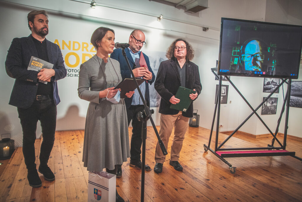 Zdjęcie. Na scenie w Galerii Winda stoją cztery osoby. Przed mikrofonem stoi kobieta w szarej sukience. Obok duży ekran, na którym widać Andrzeja Borysa.
