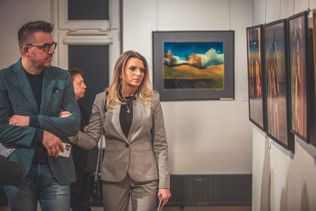 Zdjęcie. Radna Joanna Winiarska idzie pod rękę z mężczyzną patrzy na ścianę, na której wiszą fotografie Andrzeja Borysa.
