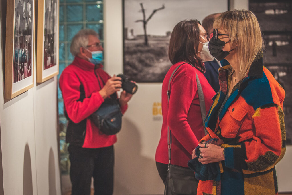 Zdjęcie. Kobieta w kolorowej grubej koszuli przygląda się fotografiom wiszącym na ścianie. W tle inni goście wernisażu oglądający prace artysty.