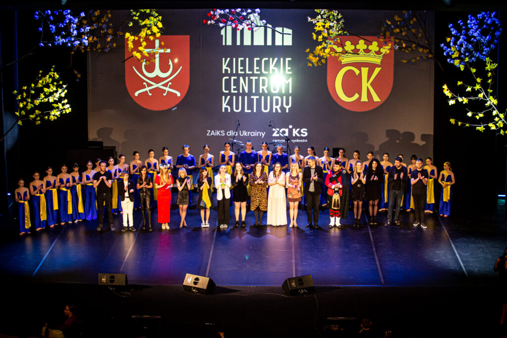 Zdjęcie. Na scenie stoją wszyscy artyści koncertu "solidarni z Ukrainą". W pierwszym rzędzie - wokaliści - w drugim tancerze w żółto-niebieskich strojach.