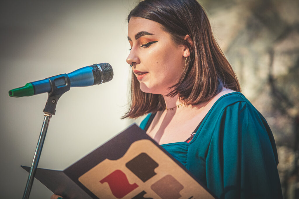 Zdjęcie. Młoda kobieta z prostymi, ciemnymi włosami do ramion, w mocnym makijażu stoi przed mikrofonem. Przed sobą ma rozłożoną dużą teczkę z literami: ZPSP