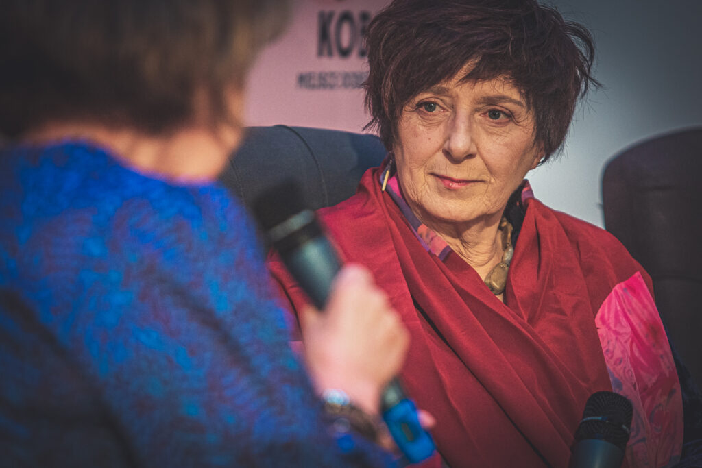 Zdjęcie. Kobieta o ciemnych krótkich włosach i asymetrycznej fryzurze patrzy w kierunku kobiety trzymającej mikrofon]