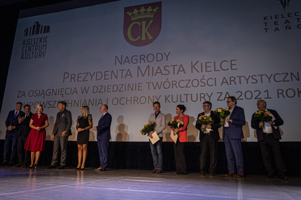 Zdjęcie. Duża Scena KCK. Na scenie ogromny ekran z napisem: nagrody prezydenta miasta Kielce za osiągnięćia w dziedzinie twórczośći artystycznej , upowszechniania i ochrony kultury za rok 2021. Przed ekranem w rzędzie po prawej i lewej stronie stoją ludzie. Po stronie prawej - przedstawiciel władz miasta i województwa. Po lewej - 6 laureatów nagród 3. stopnia, w tym Robert Wieczorek i Andrzej Skorupski..