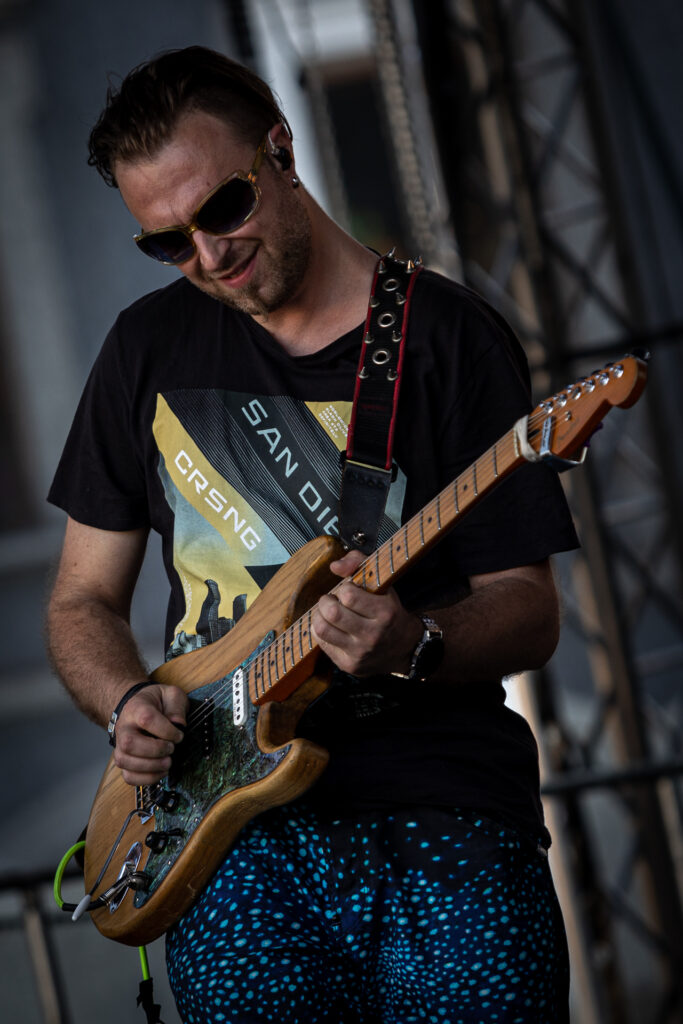 Zdjęcie. Gitarzysta w spodniach w błękitne kropki gra na gitarze. Uśmiecha się.