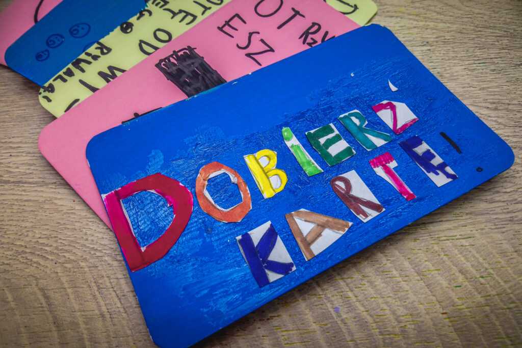 Zdjęcie. Zbliżenie na duże karty do gry przygotowane przez dzieci. Na pierwszym planie szafirowa karta z napisem: dobierz kartę!