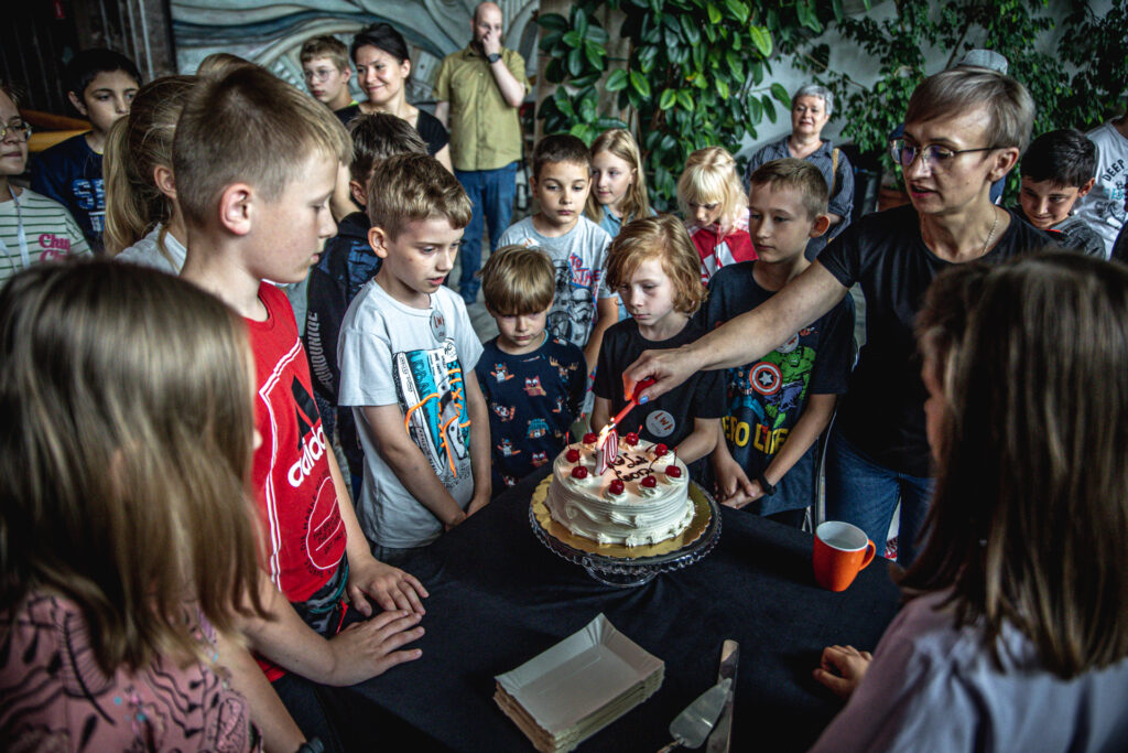 Zdjęcie. Blondynka o krótkich włosach podpala cyferki 10 na torcie. Dzieci z Lata w Teatrze stoją dookoła stolika.