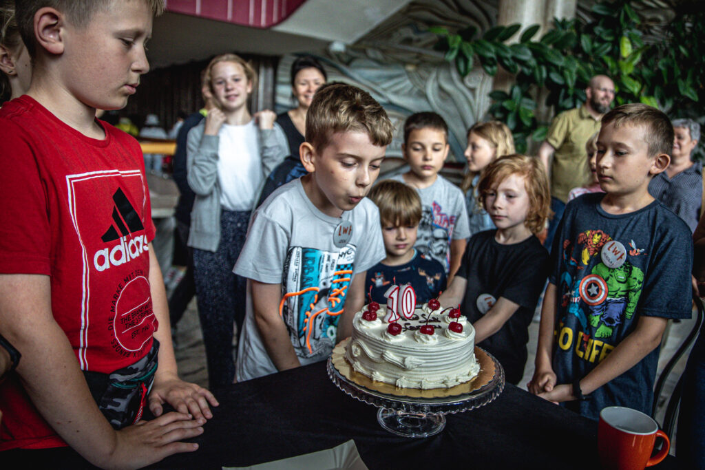 Zdjęcie. Chłopiec dmucha tort z cyframi 10 we foyer Kieleckiego Centrum Kultury. Dookoła stoją inne dzieci z Lata w Teatrze.