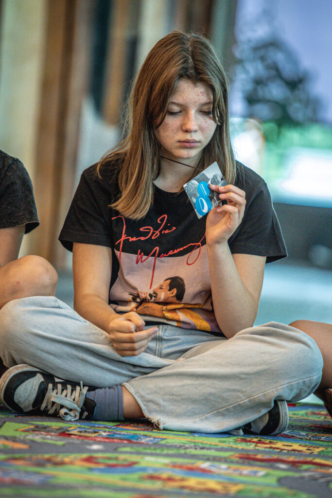 Zdjęcie. Dziewczyna siedzi po turecku. W ręku trzyma kartę do gry. Przygląda się jej.