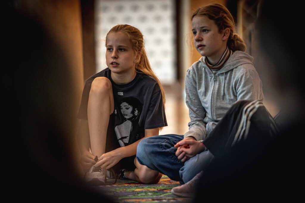 Zdjęcie. Dwie uczestniczki lata w teatrze. Po lewej: dziewczyna o długich blond włosach spiętych w kucyk i czarnej bluzce na nadrukiem zdjęcia Ariany Grande. Po prawej - dziewczyna o długich blond włosach spiętych w koczek w szarej bluzie z kapturem zapiętej pod szyję.