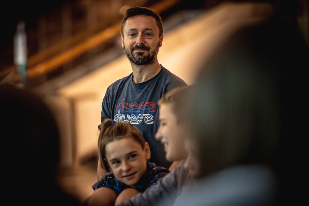 Zdjęcie. Prowadzący Michał Olszewski patrzy na grupę siedzących dzieci. Jest szczupłym mężczyzną z brodą i wąsami. Na sobie ma granatową podkoszulkę.