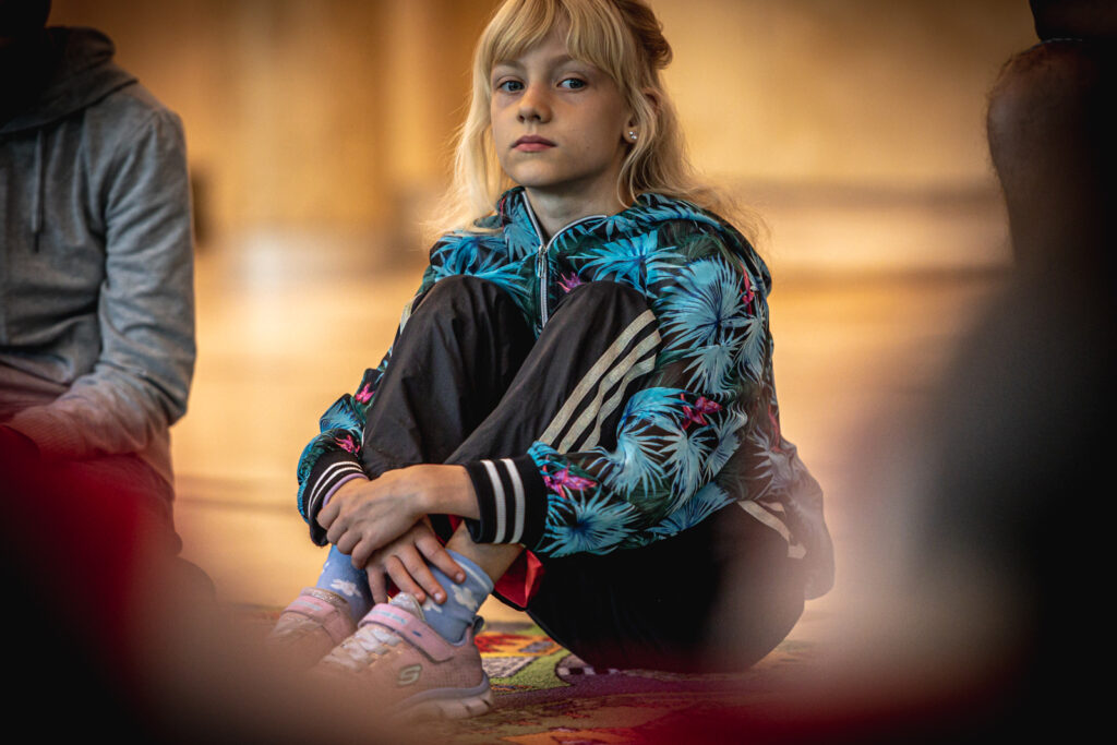 Zdjęcie. Blondynka z grzywką we wzorzystej, w turkusowe liście palmy bluzie obejmuje siedzi na wykladzinie. Ma kolana podciągnięte pod brodę. Rękami obejmuje swoje kostki.