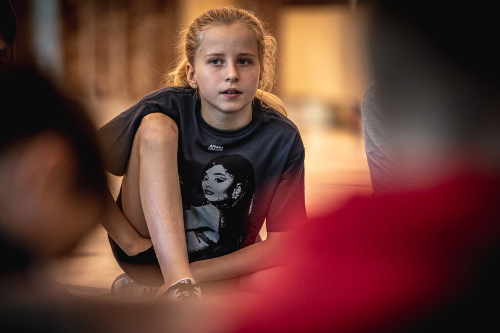 Zdjęcie. Dziewczyna o długich blond włosach patrzy przed siebie. Prawą rękę chowa pod zgiętą prawą nogą. Lewą nogę ma podsuniętą pod siebie.