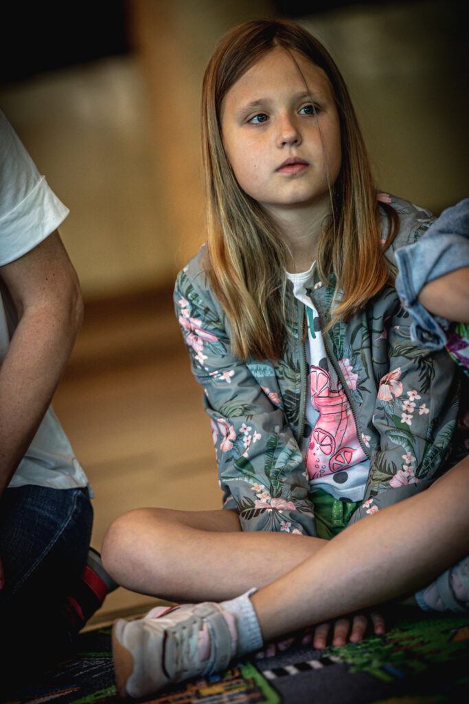 Zdjęcie. Blondynka o długich prostych włosach w kurtce w kwiaty i bluzce z różowym wzorem patrzy przed siebie.