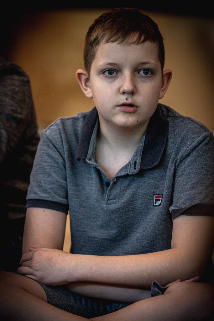 Zdjęcie. Młody chlopiec, uczestnik warsztatów ma ciemne włosy, odstające uszy i szarą koszulkę polo z logo Fila.