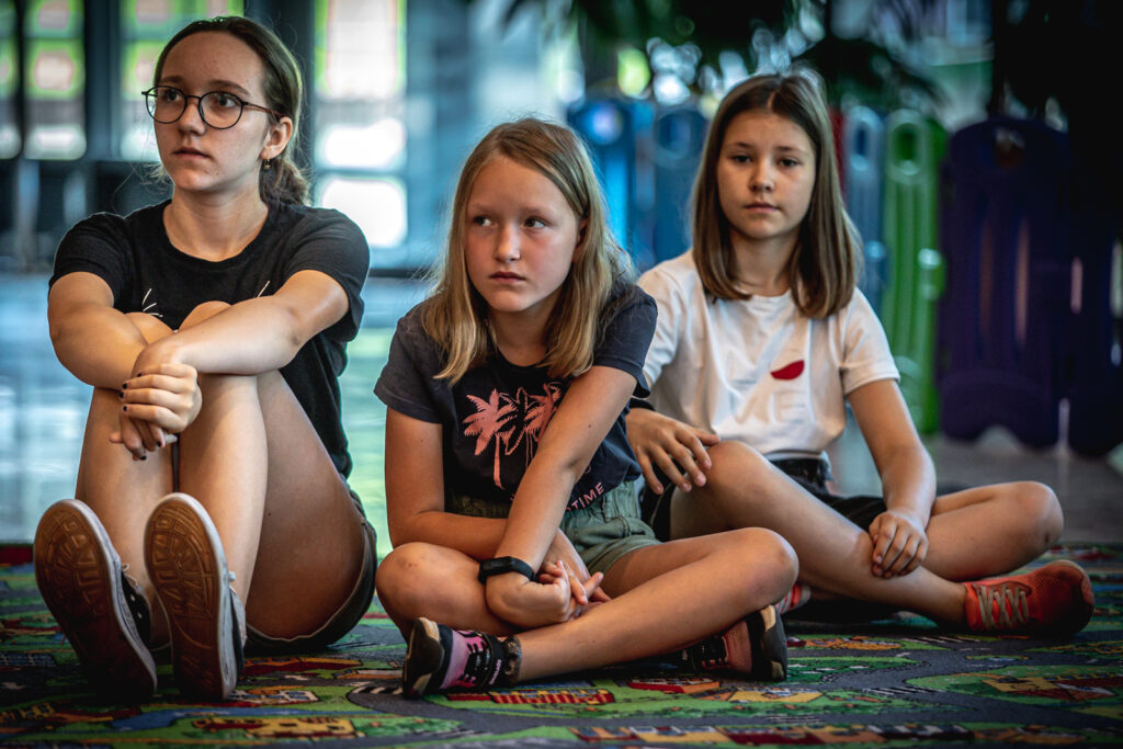 Zdjęcie. Trzy dziewczynki siedzą na wykładzinie. Najstarsza ma ciemną koszulkę, okulary na nosie i włosy spięte w kucyk. Siedzi najbardziej po lewej stronie. W centralnym miejscu ciemna blondynka w granatowej podkoszulce w różowe wzory. Ostatnia - nieco z tyłu to brunetka o długich prostych włosach poza ramiona w białej podkoszulce. To uczestniczki warsztatów Lato w Teatrze.