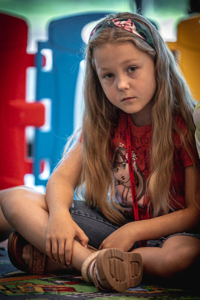Zdjęcie. Smutna blondynka o prostych, rozpuszczonych włosach z kolorową materiałową opaską siedzi po turecku na wykładzinie.
