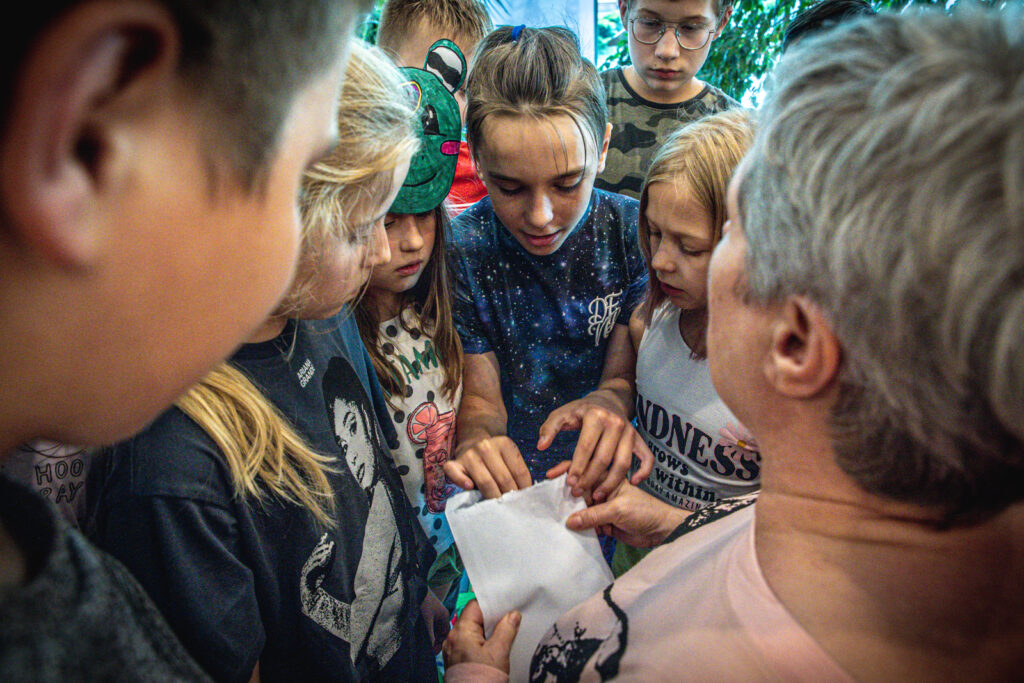Zdjęcie. Duża grupa ściśniętych dzieci. Sięgają do białej koperty.
