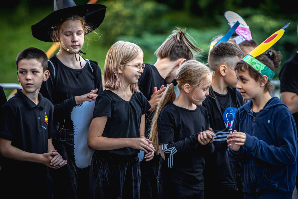 Zdjęcie. Grupa uczestników Lata w teatrze stoi na letniej scenie KCK w rzędach. Jedna z dziewczynek stoi przodem do nich.