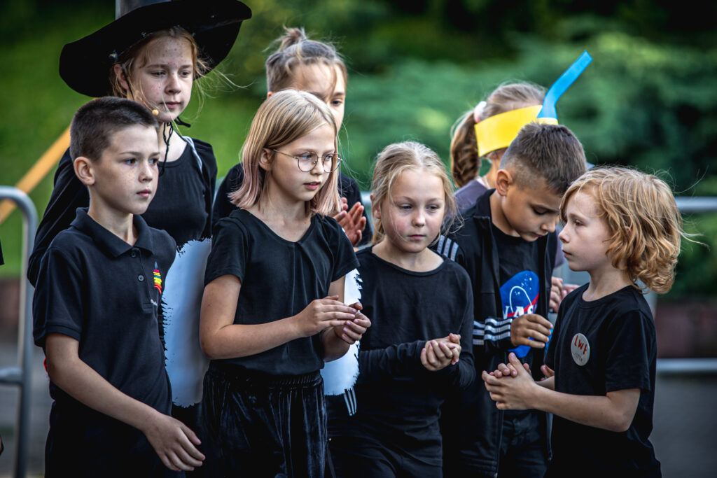 Zdjęcie. Grupa dzieci stoi blisko siebie. Przodem do nich stoi mały chłopiec o rudych włosach do ramion.