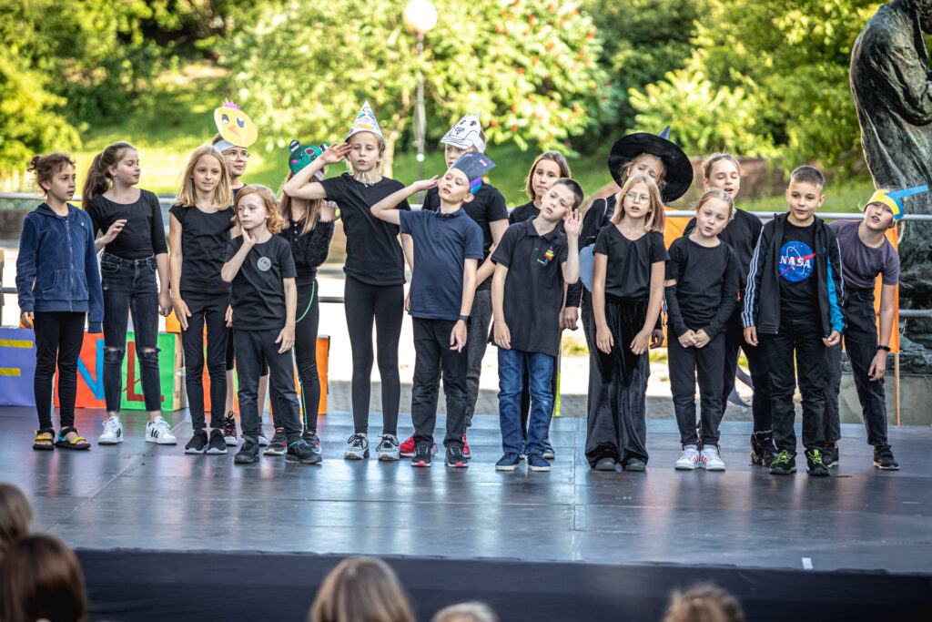 Zdjęcie. Uczestnicy lata w teatrze w śmiesznych, przygotowanych przez siebie nakryciach głowystoją w dwóch rzędach na Letniej Scenie KCK. Wszyscy pochylają się na lewą stronę i łapią się prawymi rękami za swoje prawe uszy.