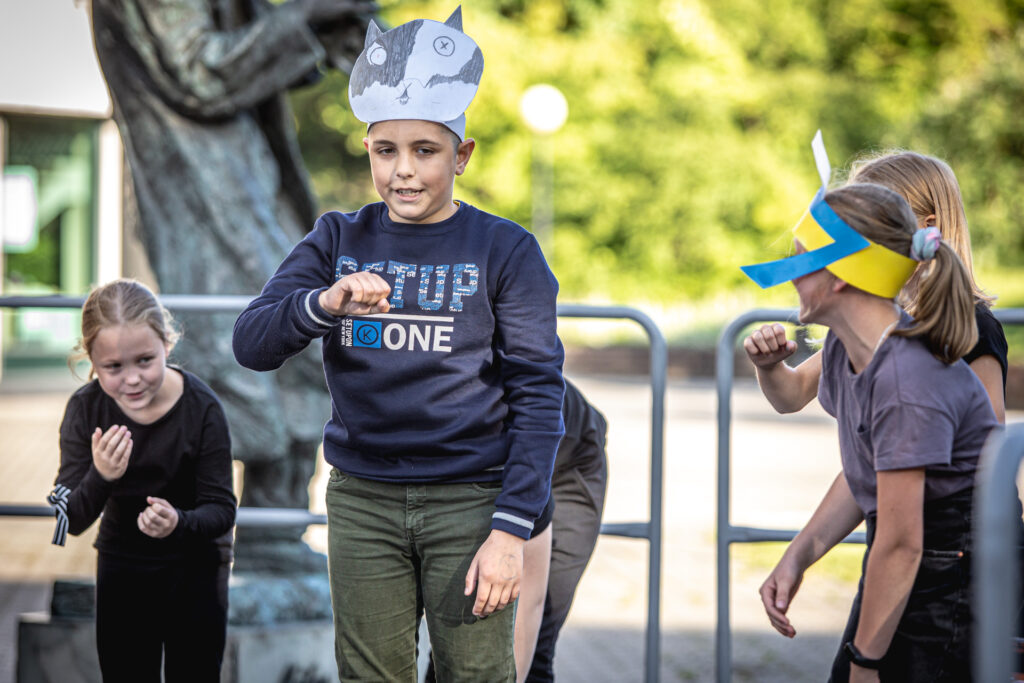 Zdjęcie. Chłopiec w granatowej bluzie z opaską w kształcie głowy czarno-białego kota. pokazuje palcem na siebie. Dookoła kilkoro dzieci stoi w półkolu, patrząc na chłopca.