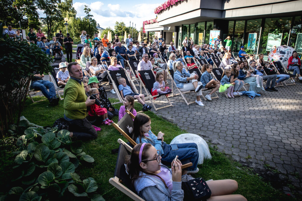 Zdjęcie. Widownia pokazu finałowego warsztatów wakacyjnych Lato w Teatrze. Ludzie siedzą na leżakach, krzesłach, i stoją na placu przed KCK.