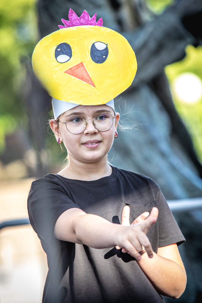 ZDjęcie. Dziewczyna w okularach z opaską z przyczepioną wielką głową żółtego kurczaka wskazuje palcem prawej ręki coś przed sobą.