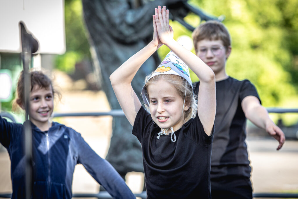 Zdjęcie. Dziewczynka o blond wlosach w szpiczastej białej czapce w kolorowe plami trzyma złączone dłonie podniesione do góry ponad głową.