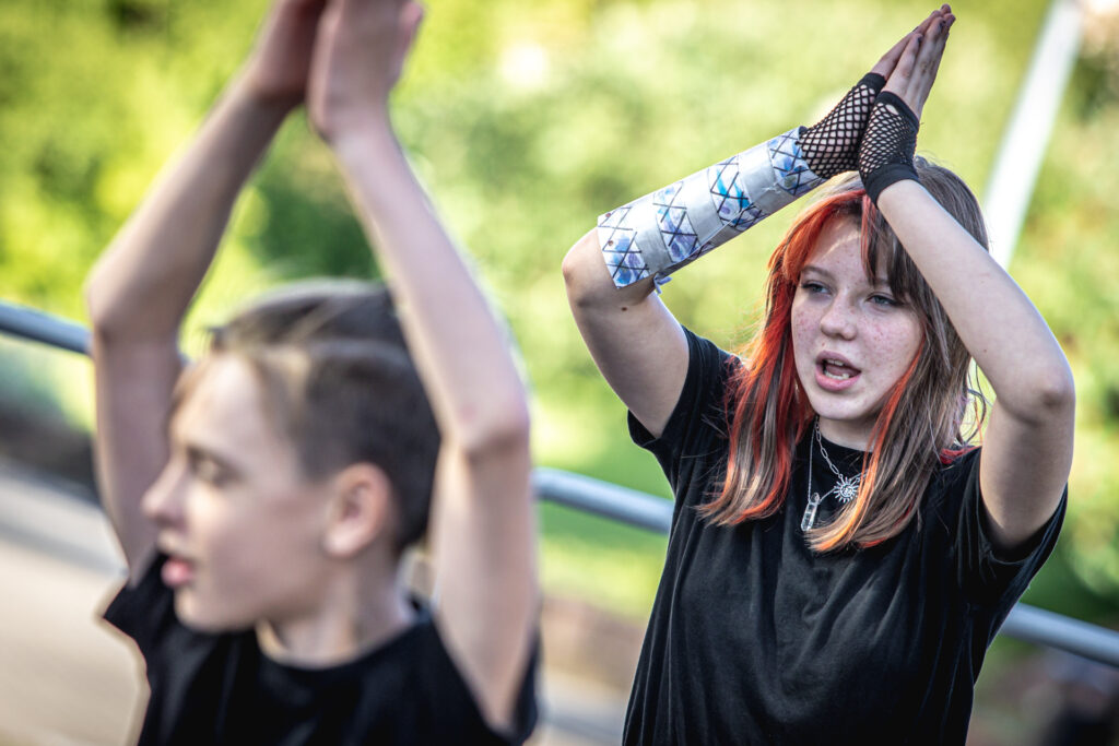 Zdjęcie. Dziewczyna z czerwonymi pasemkami na włosach. Trzyma ręce złożone razem jak do modlitwy nad głową. Przed nią chłopak robi to samo.