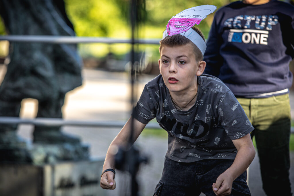 Zdjęcie. Młody chłopak z opaską z przypiętą kartonową głową przypominająca Roberta Lewandowskiego stoi na ugiętych nogach. Patrzy przed siebie z ostrym wyrazem twarz. Ręce zgięte w łokciach, dłonie zaciśnięte w pięść.