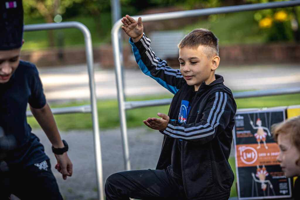 Zdjęcie. Chłopiec w ciemnej bluzie z trzema niebieskimi paskami podnosi prawą rękę do góry. Lewą wyciąga przed siebie.