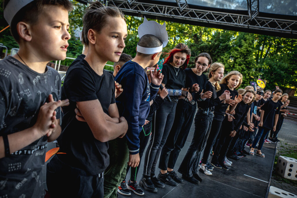 Zdjęcie. Duża grupa dzieci stoi w rzędzie na letniej scenie KCK. Jedno z dzieci trzyma mikrofon.