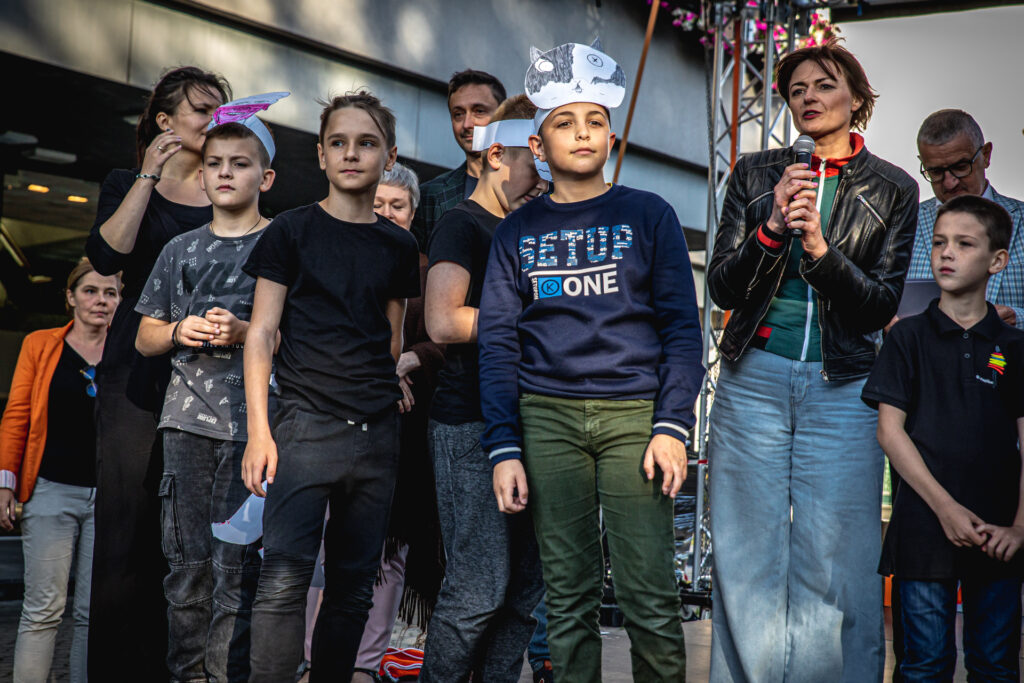 Zdjęcie. Grupa dzieci stoi na scenie. Obok kobieta zmówi do mikrofonu. To jedna z prowadzących zajęcia, Ewa Lubacz.