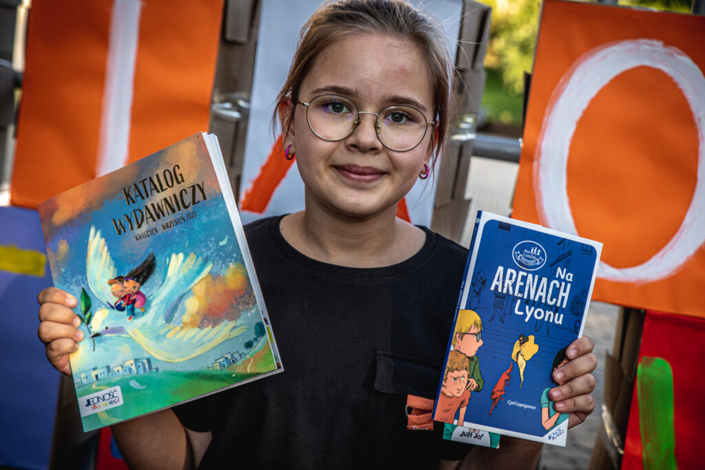 Zdjęcie. Uśmiechnięta dziewczyna w okularach pokazuje książki. Katalog wydawniczy wydawnictwa Jedność oraz Na arenach Lyonu.