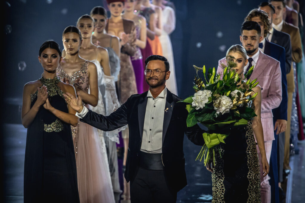 Zdjęcie. DR Paweł Węgrzyn z uśmiechem podnosi do góry ręce. W tle dwa rzędy modeli i modelek. To zakończenie jego autorskiego pokazu podczas 23. edycji OFF Fashion.