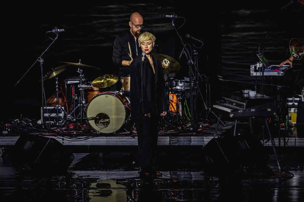 Zdjęcie. Ubrana na czarno blondynka o krotkich blond wlosach stoi przed mikrofonem. Za nią perkusista przy instrumencie. To Natalia Przybysz podczas koncertu.