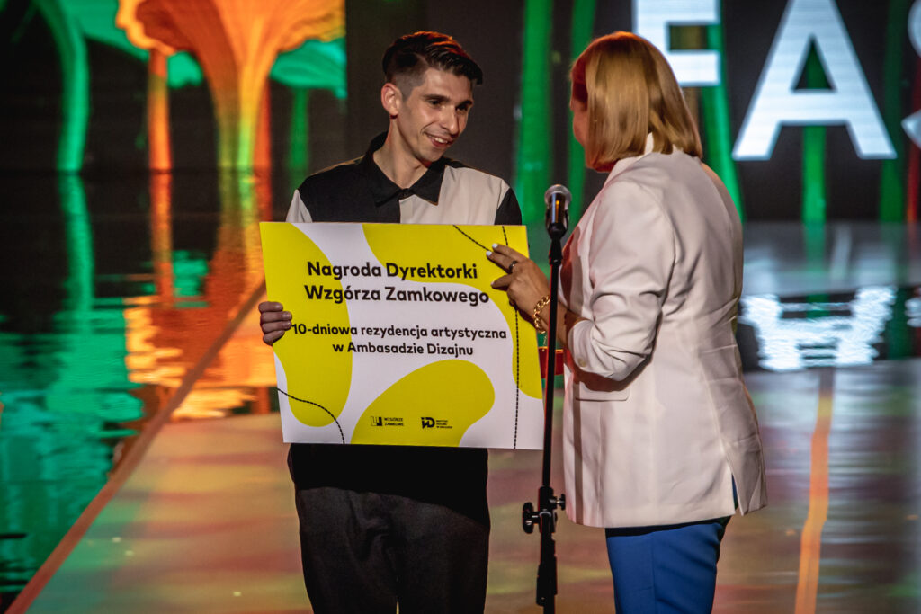 Zdjęcie. dyrektor wzgórza Zamkowego Ewa Chabielska wręcza nagrodę - 10-dniową rezydencję w Ambasadzie Dizajnu projektantowi w koszuli w połowie czarnej, w połowie białej.