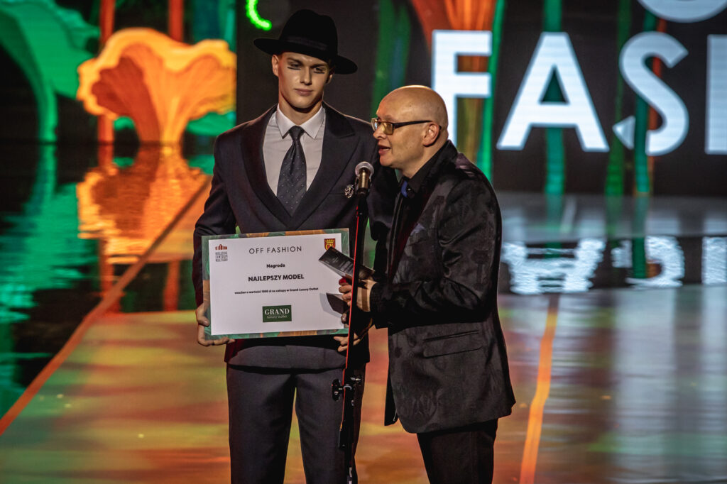 Zdjęcie. Łysy mężczyzna w okularach wręcza mówi do mikrofonu. ZA nim stoi wysoki model w czarnym kapeluszu z tabliczką dla najlepszego modela.