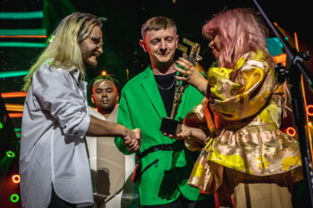 Zdjęcie. Mężczyzna w zielonej marynarce ściska rękę projektnatnowi w jasnej koszuli z długimi blond włosami. Obok niego stoi konbieta o rózowych włosach i złocistej bluzce, która w ręku trzyma statuetkę konkursu OFF.