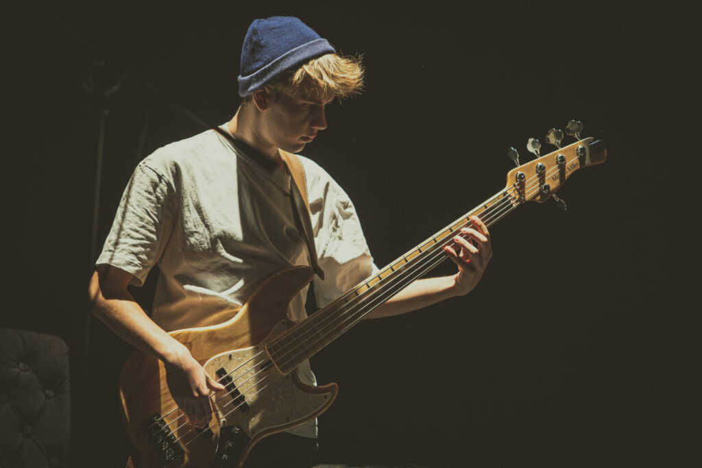 Zdjęcie. Mężczyzna z długą blond grzywką wyglądającą spod granatowej cienkiej czapki gra na gitarze.