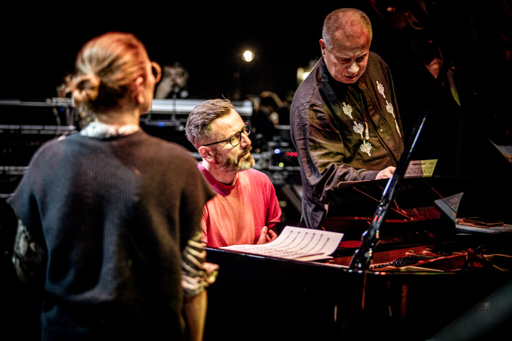 Zdjęcie. Mężczyzna w ciemnej koszuli zagląda w buty. Obok siedzi pianista w czerwonej podkoszulce. Tyłem do obiektywu stoi Anna Maria Jopek.