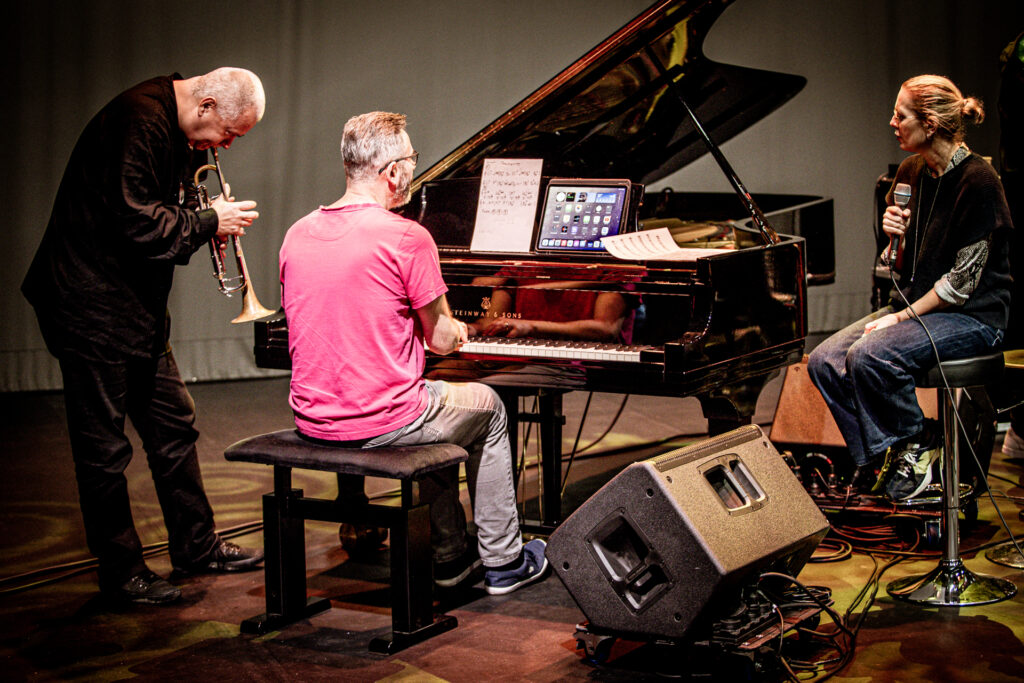 Zdjęcie. Trójka artystów na scenie. Pianista tyłem do obiektywu siedzi przy fortepianie. Po prawej - siedząca na wysokim krześle barowym blondynka z mikrofonem. Po lewej - muzyk grający na trąbce.