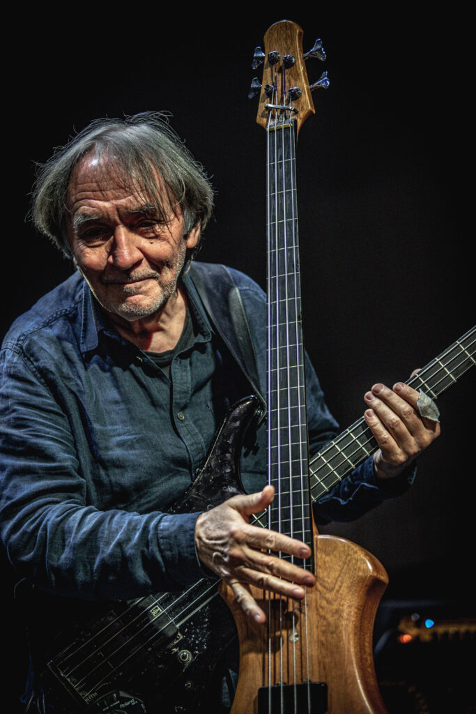 Zdjęcie. Starszy męćzyzna trzyma prawą ręką postawioną gitarę. Lewą rękę trzyma na gryfie kolejnej gitary, zawieszonej przez ramię.